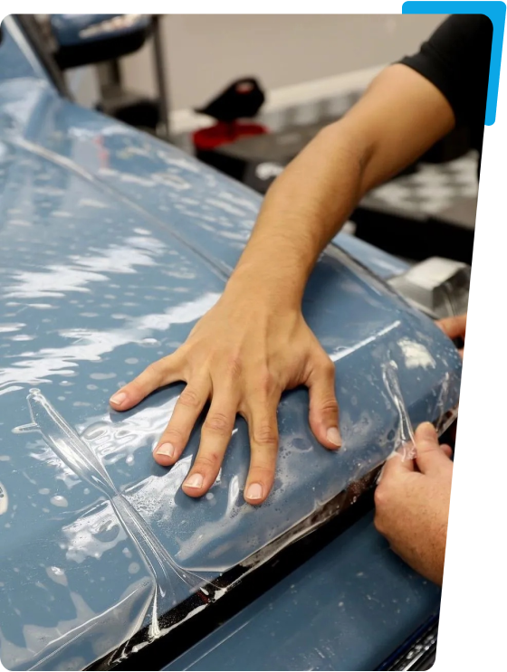 Hand applying protective film to a blue car hood, demonstrating vehicle enhancement service related to window tinting and protection.