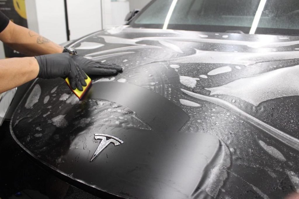 Person applying XPEL paint protection film to a Tesla hood, demonstrating professional installation for car exterior protection against damage.