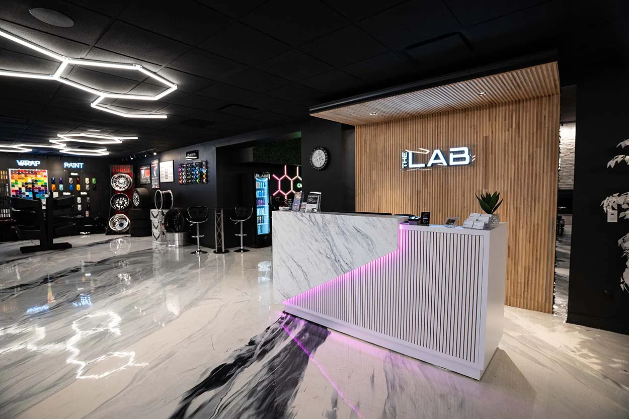 Modern auto detailing shop interior featuring a sleek reception desk with "THE LAB" signage, stylish lighting, and decorative elements, emphasizing quality and craftsmanship in a meticulously designed environment.