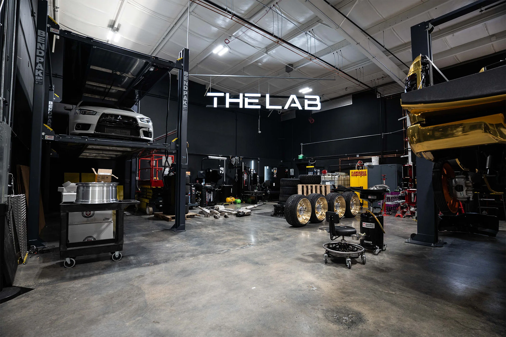 Auto detailing shop interior showcasing "THE LAB" sign, elevated vehicle lift, polished wheels, and organized workspace with tools and equipment, reflecting Aesthetics Auto Solutions' commitment to quality and meticulous design.