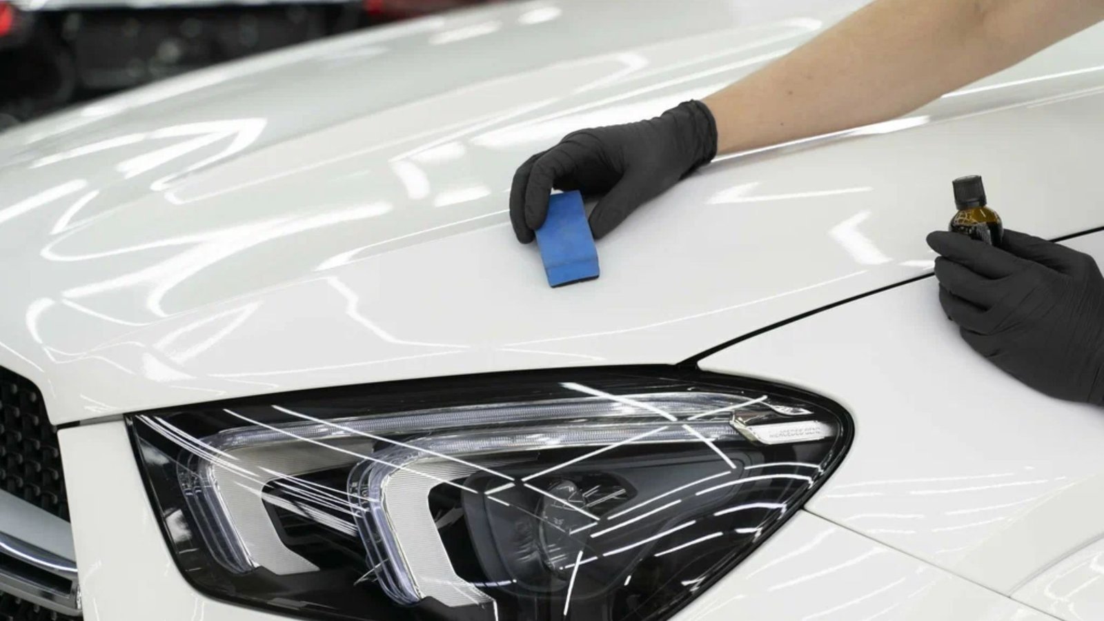 Hand applying ceramic coating to a white vehicle's hood with a blue applicator pad and a bottle of ceramic solution, showcasing automotive detailing services.