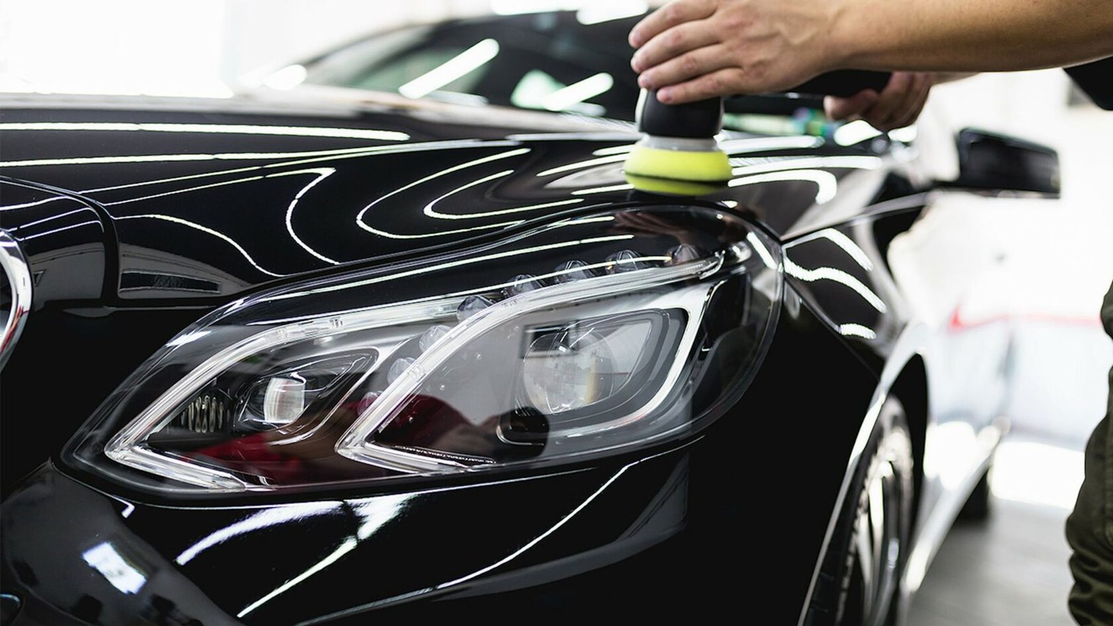 Car detailing process with a focus on polishing the headlight of a black vehicle, showcasing a technician's hand using a polishing tool in a well-lit garage setting.