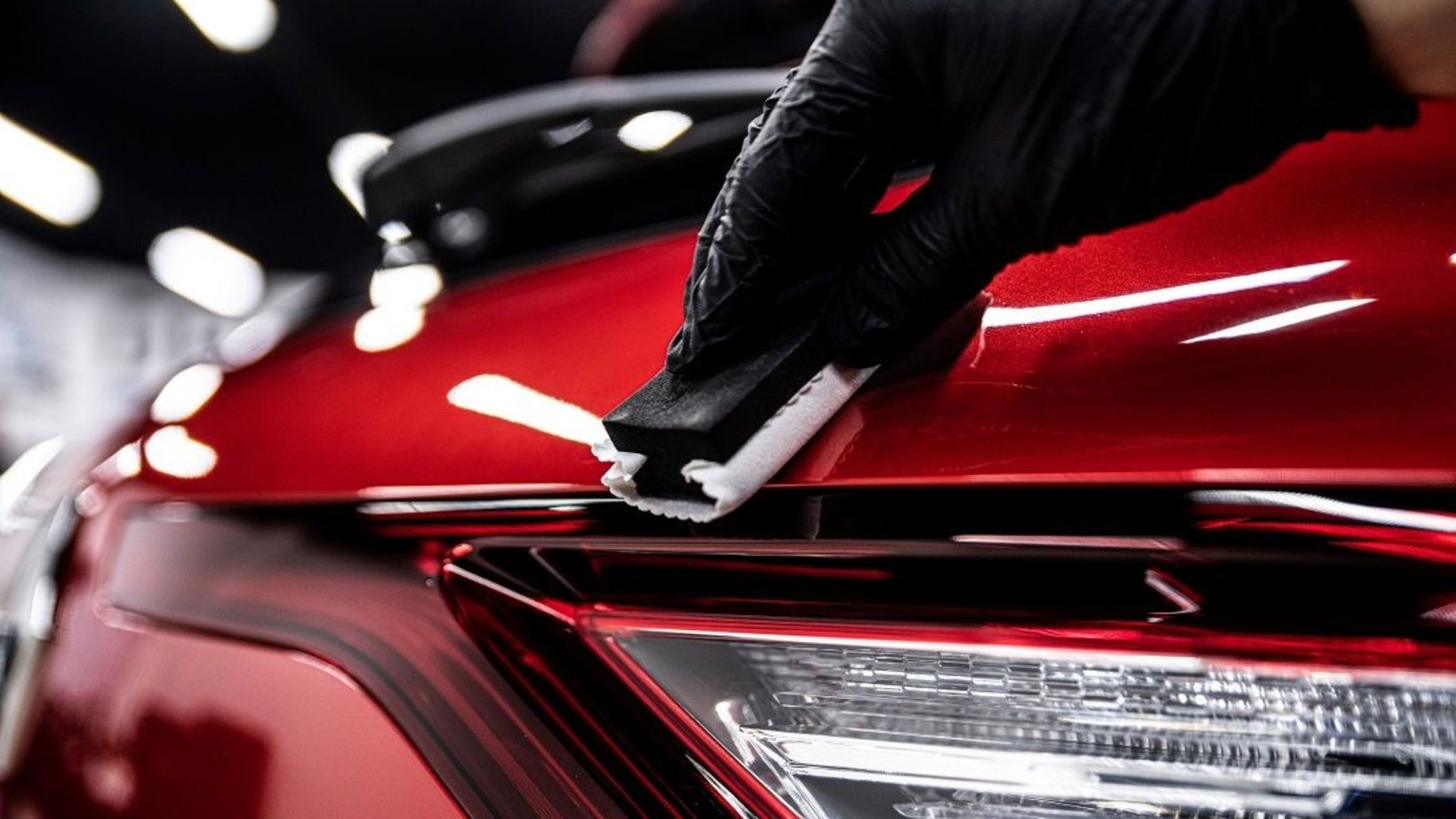 Person applying detailing product on a red car's surface, emphasizing professional car care in a detailing service environment.