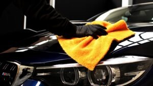 Person using an orange microfiber cloth to detail the hood of a dark blue car, highlighting professional auto detailing services offered by Aesthetic Auto Solutions in West Boylston.