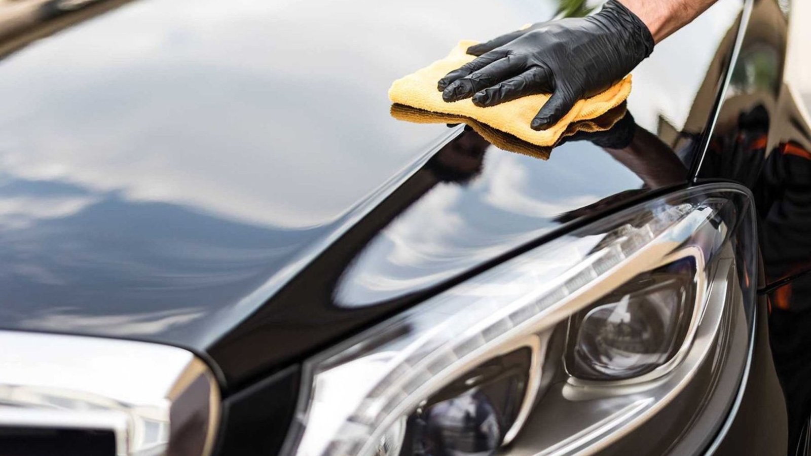 Person in black gloves polishing a shiny black car hood with a microfiber cloth, emphasizing auto detailing services for enhancing vehicle appearance and resale value.